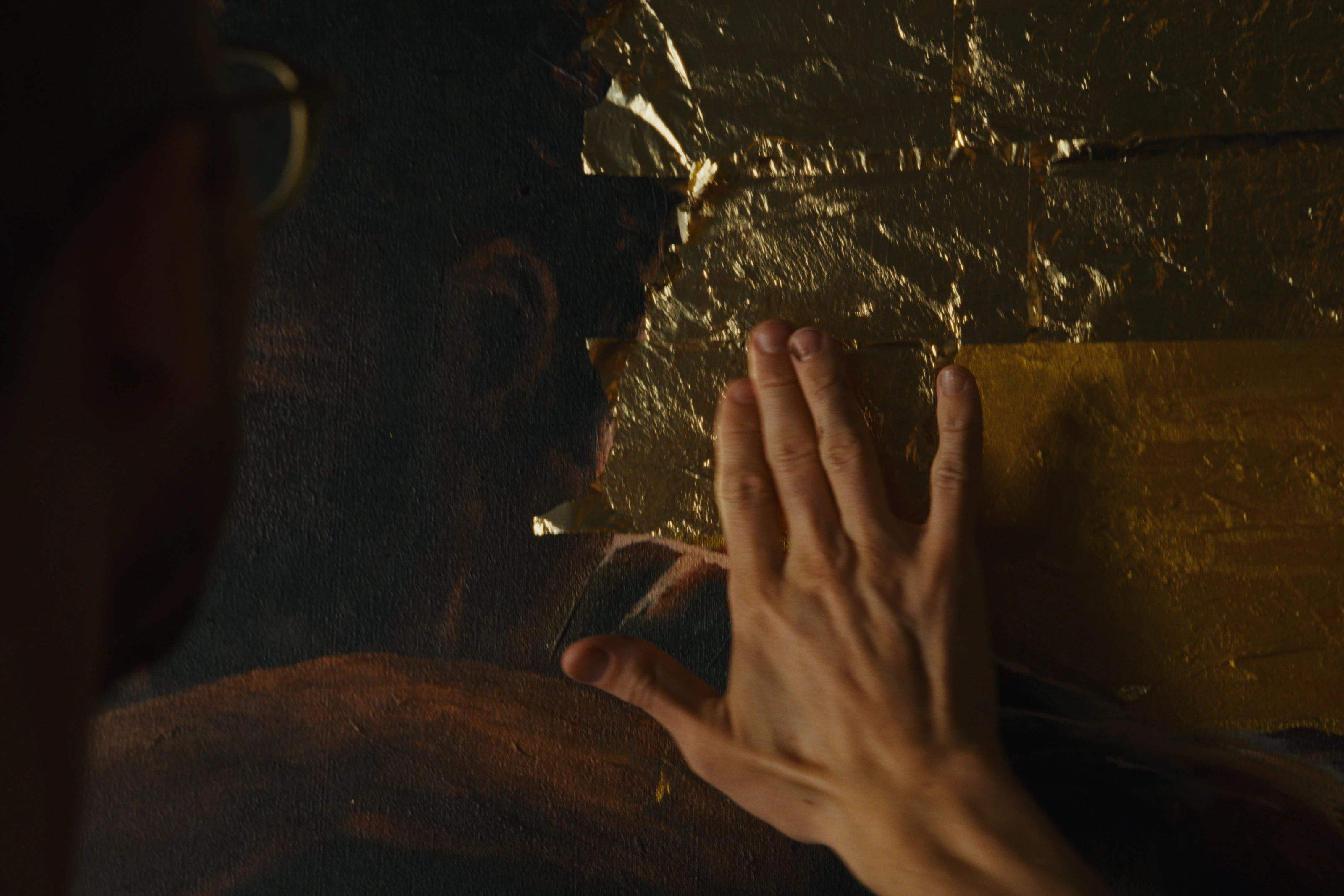 A hand touching gold leaf in a dark mural detail.